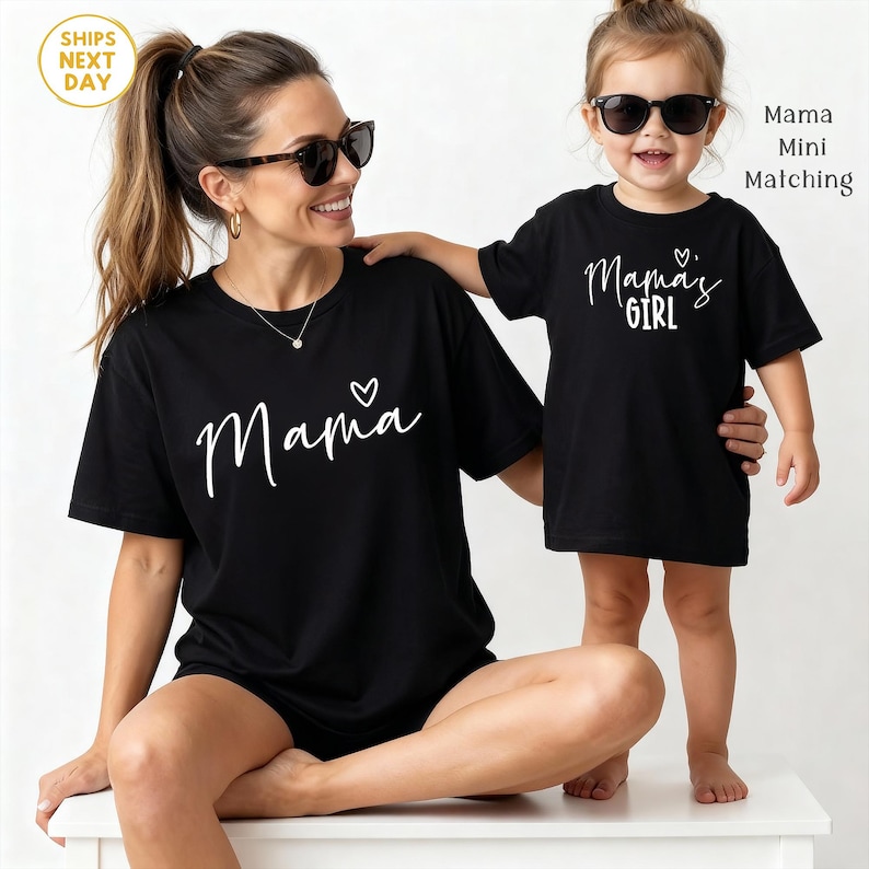 May include: Black t-shirts with white script. One shirt says "Mama" with a heart, the other says "Mama's Girl". The shirts are worn by a woman and a young child, both wearing sunglasses. The theme is matching family outfits.