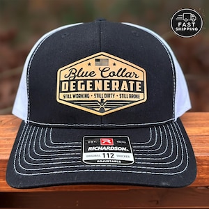 May include: Black and white trucker hat with the text "Blue Collar Degenerate" on a patch. The hat has a black front panel, white mesh back, and a curved brim with white stitching. The patch includes an American flag and the words "Still Working, Still Dirty, Still Broke."