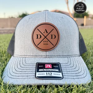 May include: A gray trucker hat with a black mesh back and a brown leather patch. The patch features the word "DAD" with "EST 2026" and crossed lines. A black and white Richardson patch is on the brim.