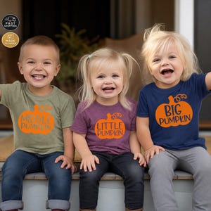 May include: Three children wearing short-sleeved t-shirts with orange pumpkin graphics. The left shirt is olive green with text reading MIDDLE PUMPKIN, the center is purple with LITTLE PUMPKIN, and the right is navy blue with BIG PUMPKIN.