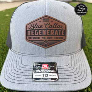 May include: A heather gray trucker hat with a black mesh back and a brown leather-like patch on the front. The patch features an American flag icon and the text: Blue Collar DEGENERATE STILL WORKING STILL DIRTY STILL BROKE. A Richardson 112 sticker is on the brim.