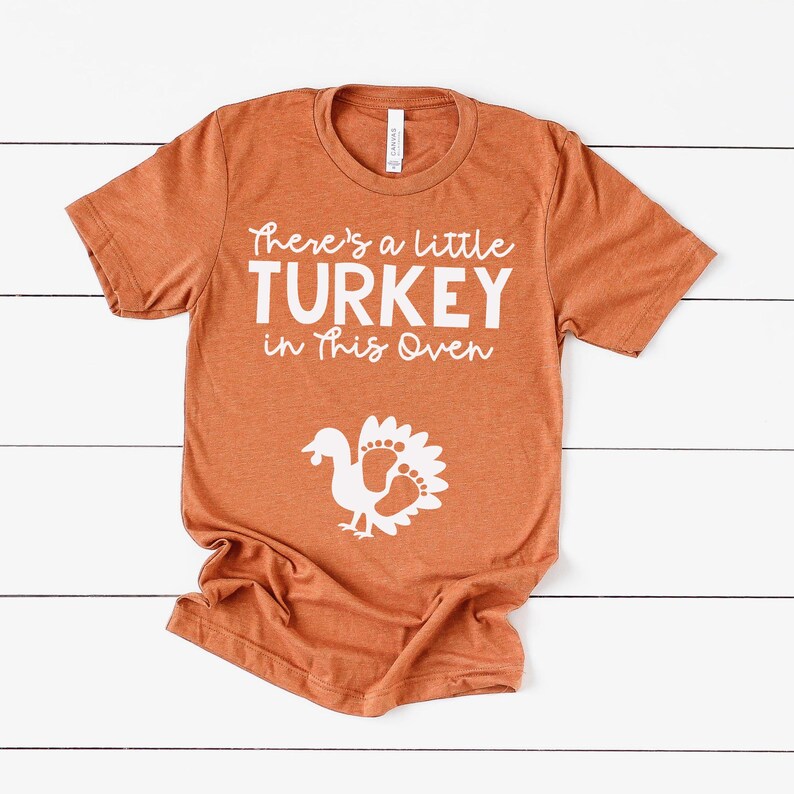 May include: A burnt orange short-sleeve t-shirt featuring white text that reads There's a little TURKEY in This Oven. Below the text is a white graphic of a turkey silhouette where the tail feathers are shaped like small baby footprints.