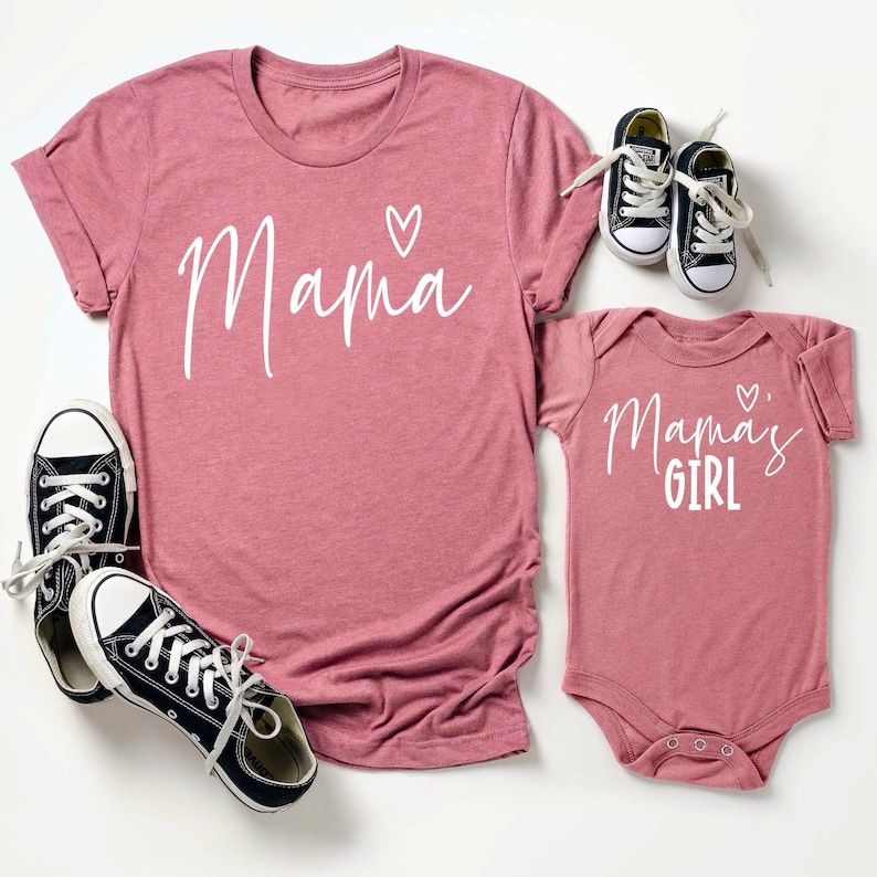May include: A matching set of mauve-colored shirts and baby bodysuit. The adult shirt says "Mama" with a heart, and the baby bodysuit says "Mama's Girl" with a heart. Black canvas sneakers are next to the adult shirt and baby shoes are next to the bodysuit.