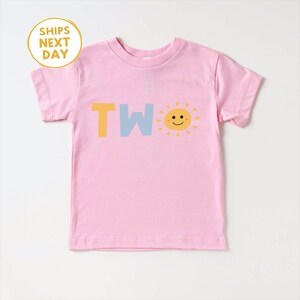 Puede incluir: Una camiseta rosa claro para ni&ntilde;os peque&ntilde;os con la palabra "TWO" en letras amarillas y azul claro, y un gr&aacute;fico de sol sonriente. La camiseta tiene mangas cortas y cuello redondo. El texto "SHIPS NEXT DAY" est&aacute; en un c&iacute;rculo amarillo.