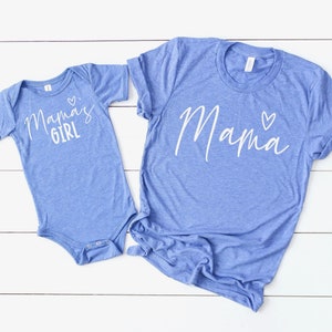 May include: Two blue t-shirts. One adult t-shirt with the word "Mama" in white script and a small heart. The other is a baby bodysuit with the words "Mama's Girl" in white script and a small heart.