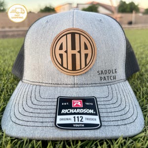May include: A gray trucker hat with a black mesh back and a curved brim. The hat features a brown leather patch with the letters "RHA" in a circular design. The text "SADDLE PATCH" is on the side. A tag reads "RICHARDSON ORIGINAL 112 TRUCKER YOUTH."