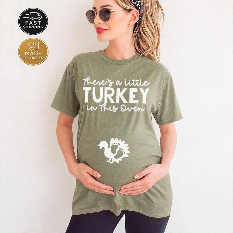 May include: An olive green short-sleeved t-shirt featuring the white text There's a little TURKEY in This Oven and a white graphic of a turkey with footprint-shaped tail feathers centered on the front of the garment.