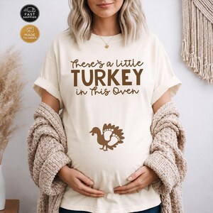 May include: A cream-colored short-sleeve t-shirt featuring the brown text "There's a little TURKEY in this Oven" printed on the chest. Below the text is a brown graphic of a turkey with footprints integrated into its tail feathers.