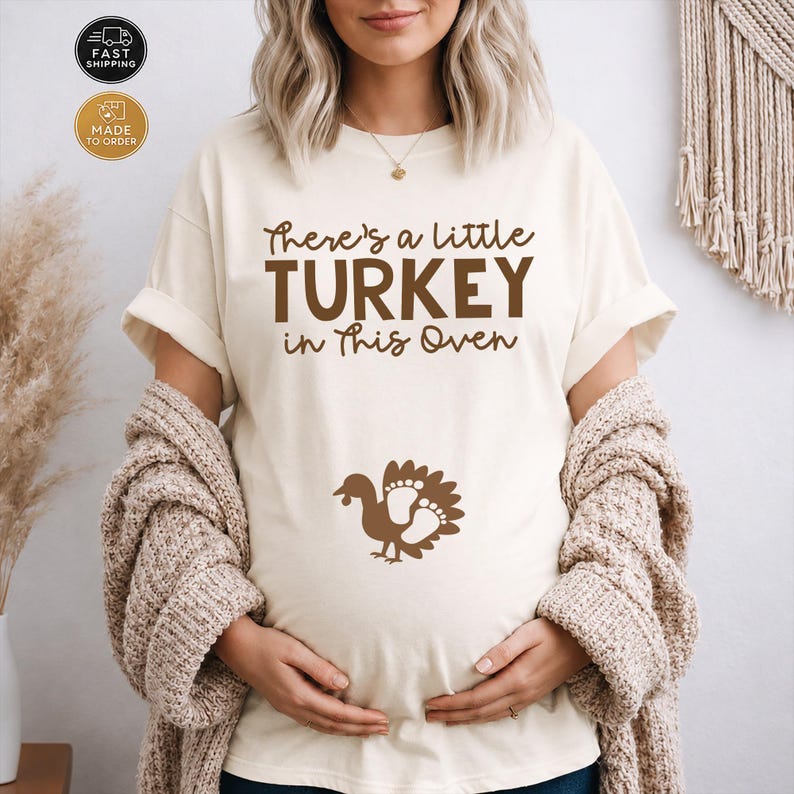 May include: A cream-colored short-sleeve t-shirt featuring the brown text "There's a little TURKEY in this Oven" printed on the chest. Below the text is a brown graphic of a turkey with footprints integrated into its tail feathers.