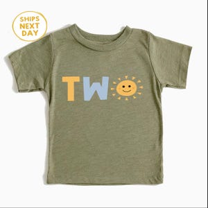 Puede incluir: Camiseta verde oliva para ni&ntilde;os peque&ntilde;os con la palabra "TWO" en amarillo y azul claro, y un gr&aacute;fico de sol sonriente. La camiseta tiene mangas cortas y cuello redondo. Un c&iacute;rculo amarillo con el texto "SHIPS NEXT DAY" est&aacute; en la esquina superior izquierda.