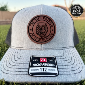 May include: A gray and black trucker hat with a leather patch that reads "I'M THE FRIEND IN LOW PLACES." The hat has a curved brim and a Richardson 112 Trucker patch. The hat is on a green grassy surface.