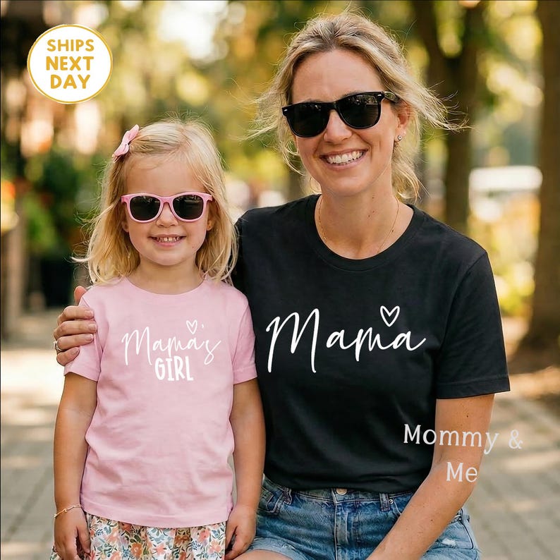 May include: A woman and a young girl wearing matching sunglasses and t-shirts. The woman's black shirt says "Mama" with a heart, and the girl's pink shirt says "Mama's Girl". The image is labeled "Mommy & Me".