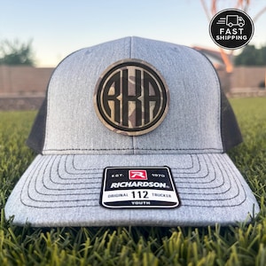 May include: A heather gray trucker hat with black mesh back panels and a circular patch featuring the letters RKA in a camouflage pattern. The brim displays a black sticker with white text reading Richardson Original 112 Trucker Youth.