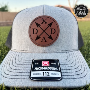 May include: A gray Richardson 112 Trucker hat with a black mesh back. The front features a brown leather patch with the letters "DAD" and crossed arrows and tools. The hat has a curved brim and a black and white tag.