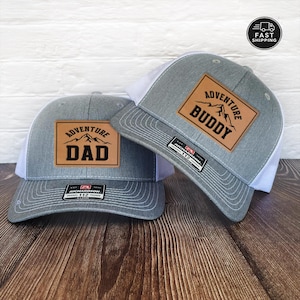 May include: Two gray trucker hats with white mesh backs and brown leather patches. One hat reads "ADVENTURE DAD" and the other reads "ADVENTURE BUDDY" in black lettering. Both hats have a Richardson 112 tag.