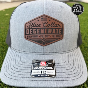 May include: Gray and black trucker hat with a brown leather patch. The patch features the text "Blue Collar DEGENERATE" and the American flag. The hat is a Richardson Original 112 Trucker, adjustable.