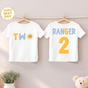 Puede incluir: Dos camisetas blancas colgadas de perchas de madera. Una camiseta tiene la palabra "TWO" en amarillo y azul con un sol sonriente. La otra camiseta dice "RANGER 2" en azul y amarillo. Ambas camisetas son para un cumplea&ntilde;os infantil.