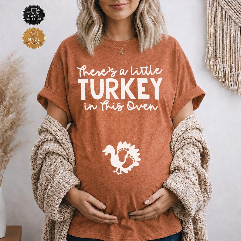 May include: A burnt orange short-sleeve t-shirt featuring the white text There's a little TURKEY in This Oven and a graphic of a turkey silhouette with footprint details. The shirt is worn with a beige textured cardigan over the shoulders.