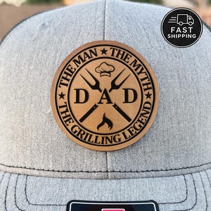 May include: A gray trucker hat with a brown leather patch. The patch features the text "DAD" in large letters, with the words "THE MAN THE MYTH THE GRILLING LEGEND" around the edge. The patch also includes images of grilling tools and a chef's hat.