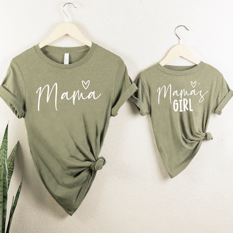 May include: Two olive green t-shirts, one adult and one child size, hanging on wooden hangers. The adult shirt says "Mama" with a heart, and the child's shirt says "Mama's Girl" with a heart. Both shirts are knotted at the bottom.