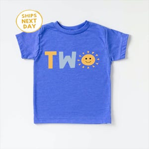 Puede incluir: Una camiseta azul para ni&ntilde;os peque&ntilde;os con la palabra "TWO" en letras amarillas y azul claro. Un gr&aacute;fico de sol sonriente est&aacute; al lado de la palabra. La camiseta es de manga corta y tiene cuello redondo. El texto "SHIPS NEXT DAY" est&aacute; en un c&iacute;rculo amarillo en la esquina superior izquierda.
