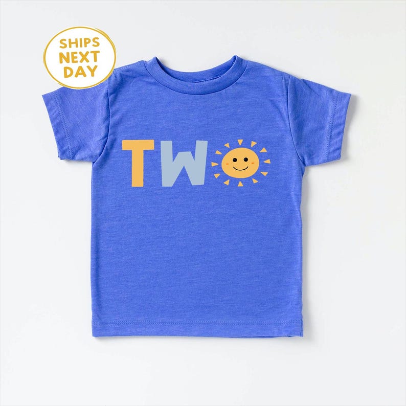 Puede incluir: Una camiseta azul para ni&ntilde;os peque&ntilde;os con la palabra "TWO" en letras amarillas y azul claro. Un gr&aacute;fico de sol sonriente est&aacute; al lado de la palabra. La camiseta es de manga corta y tiene cuello redondo. El texto "SHIPS NEXT DAY" est&aacute; en un c&iacute;rculo amarillo en la esquina superior izquierda.