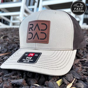 May include: Tan and brown baseball cap with a mesh back. The front features a brown square patch with the text "RAD DAD" in black. The brim is curved and has stitched detailing. A tag reads "112 The Original MD-LG".