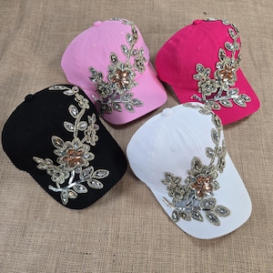 May include: Four baseball caps with floral embellishments. The caps are in black, white, pink, and red. The embellishments are made of gold and silver beads and sequins.