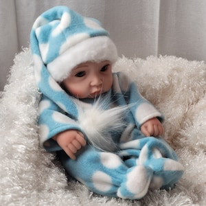 May include: A blue and white polka dot baby doll wearing a matching hat with a white fur trim. The doll is sitting on a white fluffy surface.