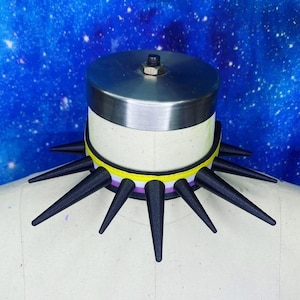 May include: A choker necklace featuring a horizontal striped ribbon in yellow, white, purple, and black. The band is adorned with a series of long, pointed black spikes extending outward around the entire circumference of the collar.