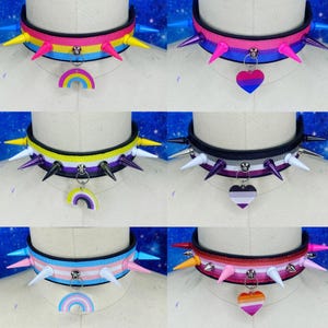 May include: A collection of six choker necklaces featuring ribbed fabric bands in various colour stripes, adorned with pointed spikes and hanging charms in either rainbow or heart shapes, displayed on white mannequin neck forms.