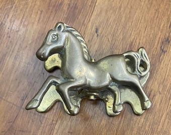 May include: A brass horse-shaped door knocker. The horse is depicted in a running pose with detailed features. The knocker has a vintage appearance and is mounted on a wooden surface.