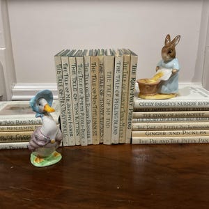 Puede incluir: Una colección de libros infantiles antiguos, incluyendo títulos como "El cuento de Peter Rabbit", y figuras de un pato y un conejo. Los libros están dispuestos sobre una superficie de madera, creando una escena nostálgica.
