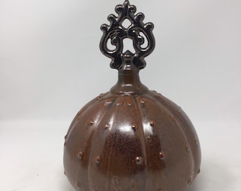 May include: A brown ceramic bottle with a decorative, dark-colored top. The bottle has a rounded shape with a textured surface and small, raised dots. The top features an ornate, swirling design.