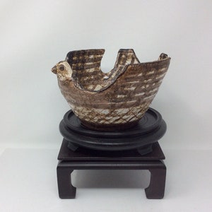 Puede incluir: Un cuenco de cerámica marrón y blanco con un diseño de pájaro. El cuenco está sobre un soporte de madera negro.