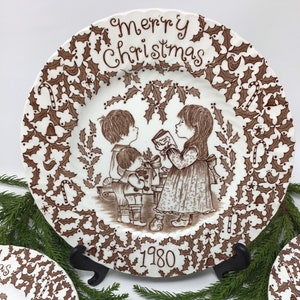 May include: A brown and white ceramic Christmas plate with a holly border and a scene of three children exchanging gifts. The plate reads "Merry Christmas 1980".