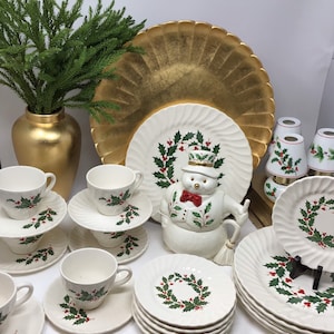 May include: A collection of Christmas-themed tableware, including white plates, saucers, and teacups adorned with holly and berry wreaths. A snowman-shaped cookie jar and a gold-colored vase with greenery are also present.