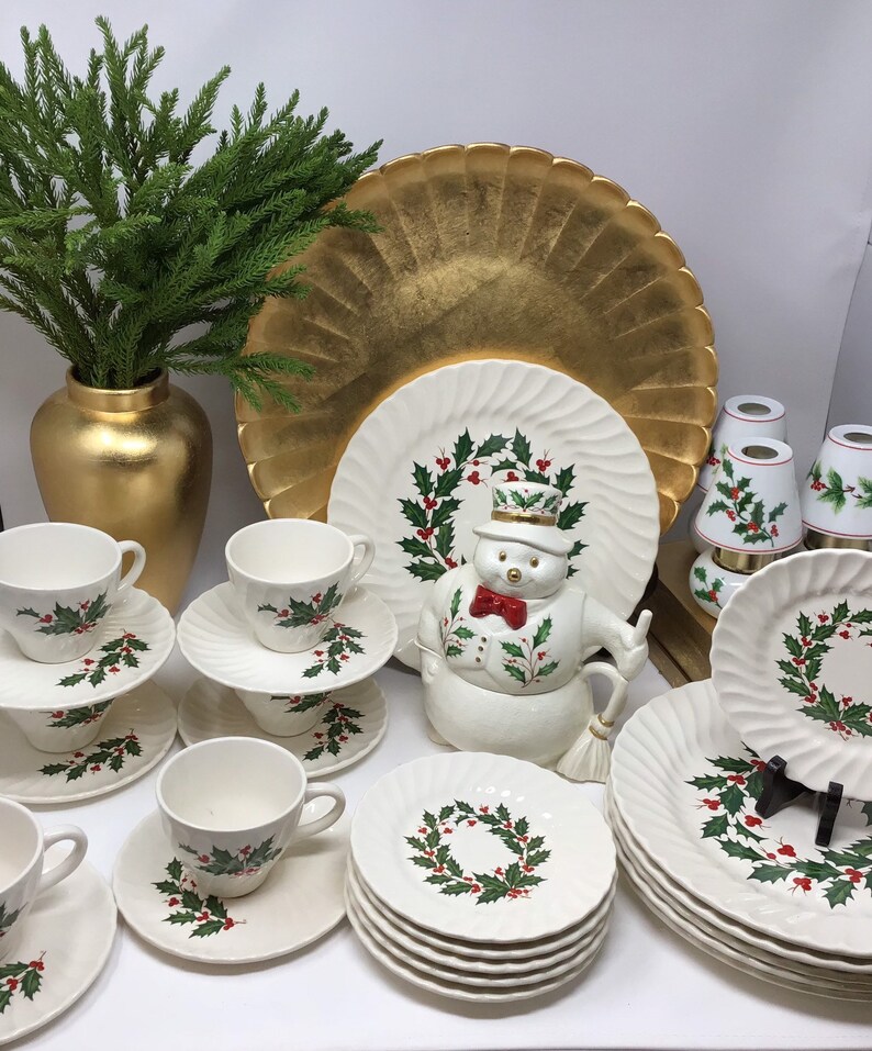 May include: A collection of Christmas-themed tableware, including white plates, saucers, and teacups adorned with holly and berry wreaths. A snowman-shaped cookie jar and a gold-colored vase with greenery are also present.