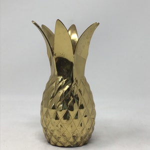 May include: A gold-colored pineapple-shaped decorative object. The pineapple has a textured, diamond-patterned body and a crown of pointed leaves. The object is made of metal and has a slightly aged appearance.