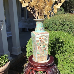 May include: A tall, rectangular, antique Chinese vase with a light turquoise background and detailed figures. The vase sits on a dark brown, carved wooden stand. The vase holds a bundle of dried, golden-colored decorative branches. The scene is outdoors.