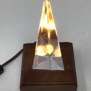 May include: A pyramid-shaped lamp made of clear glass, emitting a warm, amber light. It is mounted on a dark brown, rectangular wooden base. A brown electrical cord is attached.