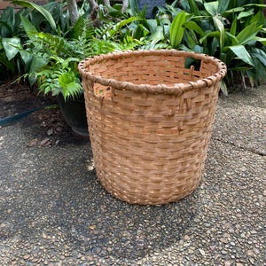 May include: A round, light brown woven basket featuring a sturdy, open-top design with a thick, braided rim. The basket is constructed from flat, interlaced wooden strips, creating a consistent grid pattern throughout the cylindrical body.