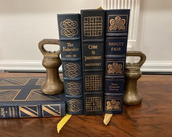 Puede incluir: Una colección de libros clásicos, incluyendo "The Federalist", "Crime & Punishment" y "Vanity Fair". Los libros están encuadernados en azul oscuro y negro con letras doradas y detalles decorativos. Dos sujetalibros antiguos de latón flanquean los libros.