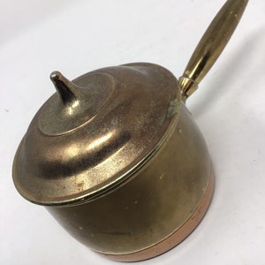 May include: A small, vintage saucepan with a lid and a long handle. The pot is made of brass and copper, with a gold-colored handle and lid. The saucepan is designed for cooking and has a classic, retro aesthetic.