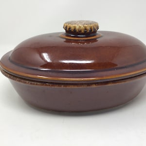 May include: A brown ceramic casserole dish with a lid. The dish is oval-shaped with a glossy finish. The lid has a small, textured handle. The dish is designed for cooking and serving food.