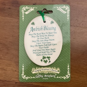 Puede incluir: Adorno ovalado de porcelana con una bendición irlandesa impresa en verde. Está unido a una tarjeta verde con bordes decorativos y el texto "Hanging Ornament Collection" y "Liffey Artefacts Ireland".