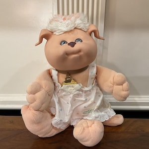 Könnte beinhalten: Eine Plüschpuppe mit einem weichen, pfirsichfarbenen Vinylkopf, hellbraunen Ohren und weißem, flauschigem Haar. Die Puppe hat blaue Augen, eine kleine braune Nase und trägt ein weißes ärmelloses Kleid mit kleinen Herzchen und einem goldenen Anhänger.