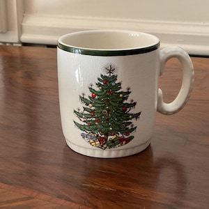 May include: White ceramic mug with a green rim and handle. The mug features a detailed illustration of a Christmas tree decorated with ornaments and presents at the base. A festive holiday-themed mug.