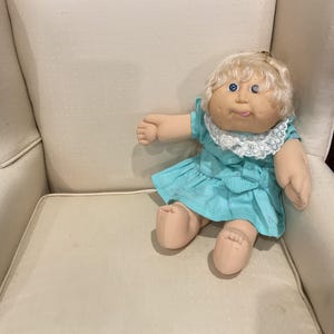 May include: A doll with blonde yarn hair, blue eyes, and a light skin tone, wearing a bright turquoise short-sleeved dress with a white lace collar. The doll has a soft fabric body, rounded facial features, and a small tongue visible at the mouth.