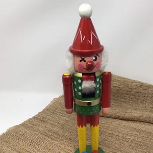 May include: A wooden nutcracker figurine dressed as a clown with a red pointed hat, white pom-pom, and white hair. The figure features a green jacket with white polka dots, yellow pants, and a round red nose, standing on a circular green base.